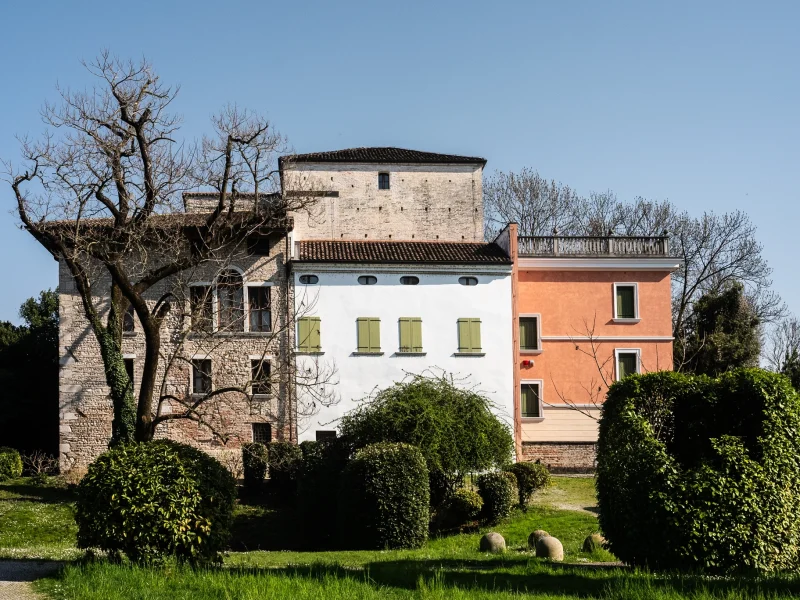 Edificio composto da tre corpi affiancati, con facciate di colori diversi: una struttura in mattoni a sinistra, una parte centrale bianca con imposte verdi e una sezione arancione a destra. Davanti all’edificio si trovano siepi potate e un prato verde. Sullo sfondo sono visibili alberi spogli e cielo sereno.