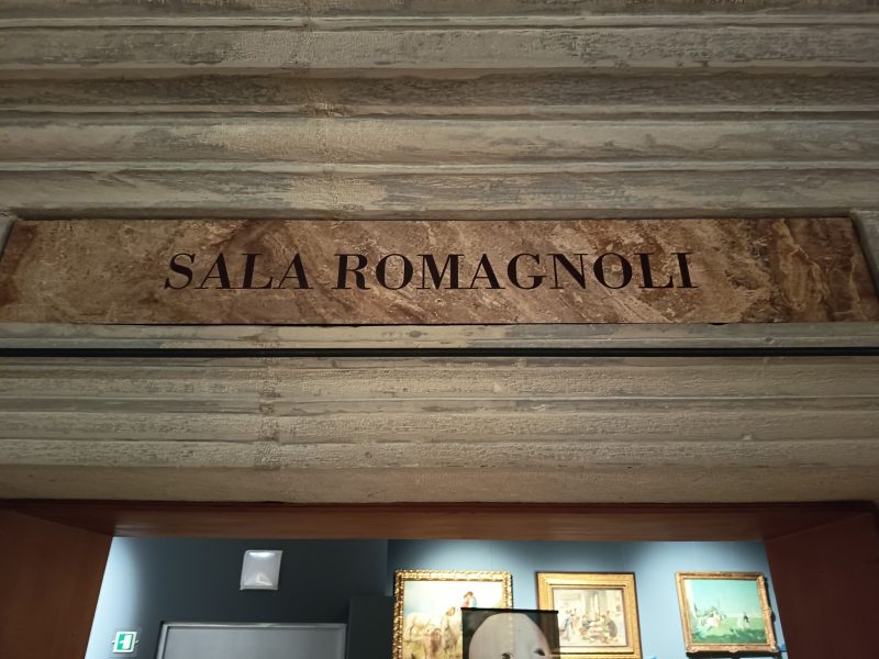 Ingresso di una sala museale con architrave in pietra recante la scritta “Sala Romagnoli”, sopra una porta che conduce a uno spazio espositivo.