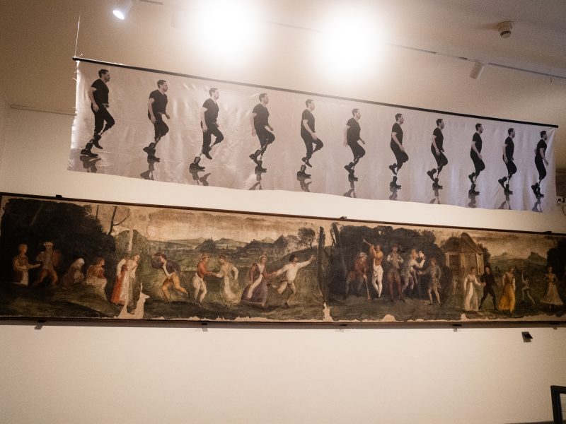 Una veduta di una parete espositiva bianca che mette a confronto due opere d'arte orizzontali e parallele, una moderna e una antica, sul tema del movimento. In alto: Uno striscione fotografico contemporaneo in bianco e nero mostra una sequenza cronofotografica di un uomo con una protesi alla gamba mentre corre. La sequenza cattura undici diversi istanti del passo, creando un senso di dinamismo fluido. In basso: Un grande affresco o fregio antico, di forma lunga e stretta, raffigura una scena conviviale e campestre. Numerose figure in abiti d'epoca sono ritratte mentre danzano in coppia o si muovono in un paesaggio rurale con alberi e colline. Lo stile è rinascimentale o barocco, con tonalità calde e terrose ormai sbiadite dal tempo. L'illuminazione proviene da faretti a soffitto che creano due grandi riflessi luminosi sopra lo striscione fotografico, enfatizzando il contrasto tra la tecnologia moderna della protesi e la storicità del dipinto sottostante.