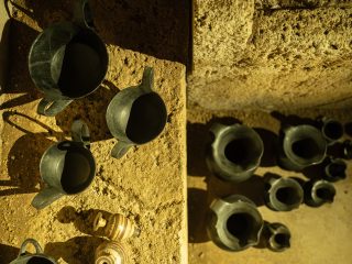 Veduta dall'alto di un gruppo di antichi vasi in ceramica nera, disposti su un pavimento di pietra grezza e illuminati da una luce calda laterale. A sinistra predominano coppe e brocche con grandi anse, mentre a destra ci sono vasi globulari e piccoli bicchieri. In basso, si intravedono due piccoli vasi con decorazioni lineari policrome. Le ombre allungate creano contrasto sulla superficie rocciosa.