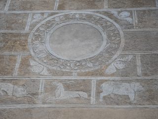 Detail of an exterior graffito on Palazzo Marone in Pordenone, depicting a geometric panel design: at the centre is a large circular medallion engraved with plant motifs and surrounded by cherub heads at the corners, whilst the lower section features animal silhouettes, including a deer and a wild boar, engraved into the mortar.