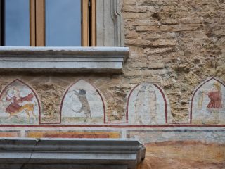 Detail of a fresco frieze painted beneath a stone cornice at Palazzo Marone in Pordenone. Four ogival panels separated by red frames follow one another on a rough-cast wall: the first depicts a female centaur with a bow and arrows, the second a climbing bear, the third two stylised figures of fish facing each other, and the fourth a human figure wearing a cap.