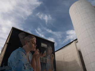 "A large mural painted on the dark wall of a building, depicting a woman applying make-up whilst looking into a small mirror. She is wearing a light blue garment decorated with bird motifs. Next to the main figure is a fainter image of a person who appears to be doing the same thing. To the right of the building stands a modern structure featuring a tall cylindrical tower clad in square white tiles, all set against a blue sky with scattered clouds."