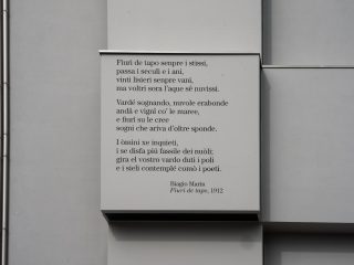 ‘A detail of a rectangular grey architectural panel protruding from a modern wall with a minimalist design. A poem in dialect is printed in black on the panel, with the attribution “Biagio Marin, Fiuri de tapo, 1912” at the bottom. To the left of the panel, two metal tubes run vertically along the surface of the wall.’