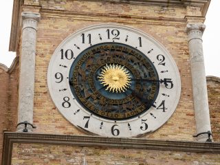 A detail of an antique astronomical clock set against an exposed brick façade, flanked by two stone columns. The main dial features a traditional Roman numeral marking from 1 to 12. In the centre, a smaller rotating disc shows a radiant golden sun surrounded by a ring bearing the signs of the zodiac and the names of the months in archaic Italian, engraved along the circumference. The dark hands indicate the time, whilst the central mechanism illustrates the apparent motion of the celestial bodies.
