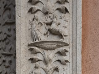 ‘A detail of a carved stone relief on a vertical pillar, divided into sections by stylised plant motifs. The carvings depict allegorical scenes or zodiac signs, including a centaur shooting an arrow (Sagittarius), a figure holding a set of scales (Libra) and other human figures, all set against a background of engraved stars.’