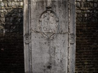 ‘An ancient stone slab set into a wall of bricks and rustic stone. In the centre, the slab features a coat of arms carved in relief at the top and a Latin inscription, partially worn away by time, which occupies the central section.’