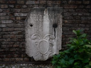 ‘A detail of a light-coloured plaster façade, showing signs of wear, featuring a painted coat of arms between two windows. The coat of arms depicts a golden lion on a grey background in the upper section and red and white horizontal bands in the lower section. On either side are two windows with green wooden shutters that are visibly weathered.’