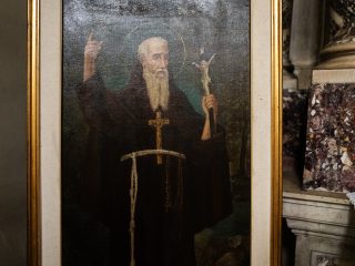 "Dipinto a olio raffigurante un santo barbuto con aureola, che indossa un saio scuro tipico degli ordini monastici, con un crocifisso al collo e un cordone annodato in vita. Il soggetto tiene la mano destra alzata con l'indice puntato verso l'alto e regge un piccolo crocifisso nella mano sinistra. Il quadro è inserito in una cornice dorata all'interno di un ambiente religioso."