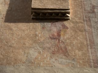 The image shows an architectural detail of an ochre/beige plaster wall. Beneath a relief cornice featuring a series of triangular mouldings (with serrated edges), the remains of a faded fresco can be glimpsed, depicting a human figure—probably a saint or an allegorical figure—clad in reddish robes.