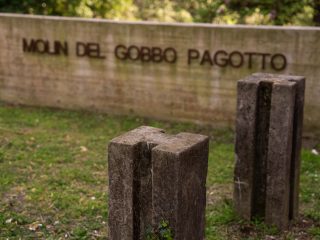 The image shows an outdoor area, probably a park or a historic site, with a concrete backdrop to which the metal letters forming the name are affixed. In the foreground are two small rectangular stone or concrete pillars, each featuring a deep vertical slit running down the centre of each side.