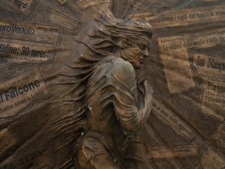 The image shows a bronze sculpture featuring a central human figure in relief, whose features and clothing appear to merge in a dynamic movement. The background of the sculpture consists of numerous panels that faithfully reproduce the headlines and articles of Italian newspapers, transforming the surface into a sort of engraved historical archive.
