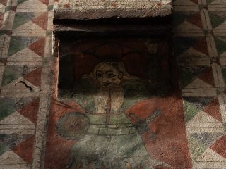 Detail of a frescoed interior wall in Palazzo Marone in Pordenone. In the centre, a bust of a medieval warrior is depicted wearing green scale armour, with a sword at his belt and a small circular shield. The warrior has a long blond beard and a fixed gaze. Above him, a section of wall featuring a geometric zigzag and diamond pattern rises like a column. The background is brick red and shows signs of wear, whilst a series of painted frames surrounds the fresco.