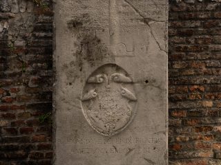 L'immagine mostra una lapide commemorativa o una pietra tombale in pietra chiara, posizionata tra due pareti di mattoni rossi. Al centro della lastra si trova un emblema araldico scolpito in bassorilievo all'interno di una forma ovale, raffigurante dei motivi floreali o animali stilizzati sormontati da una corona.