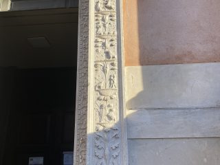 Detail of a carved stone jamb from a monumental portal, richly decorated with Renaissance-style bas-reliefs featuring candelabra motifs. The decoration alternates plant motifs, such as vases and acanthus leaves, with small human and animal figures arranged in vertical panels. The light-coloured stone pillar contrasts with the adjacent antique pink wall, partially illuminated by side sunlight which enhances its three-dimensionality. To the left, one can glimpse the dark entrance to a sacred building with several notices posted on the door.