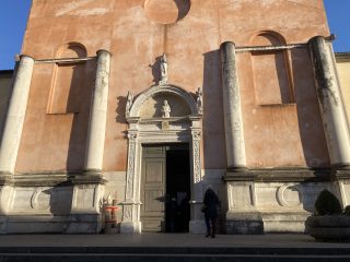 The façade of a historic church, characterised by pale pink plasterwork. The main entrance is framed by a carved stone portal, surmounted by a sculpted lunette depicting Christ, and crowned by a statue in an upper niche. Two large stone columns appear to support the central section of the façade, which features empty side niches. A person stands in the doorway, ready to enter. On either side of the portal are sculptural decorations and vases with plants. Sunlight casts sharp shadows on the structure, highlighting its architectural forms.