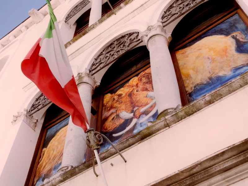 Inquadratura dal basso della facciata esterna principale del Museo di Storia Naturale di Pordenone. In una delle trifore del palazzo è inserito un dipinto che raffigura due mammut duellanti. In primo piano a sinistra, una bandiera italiana sventola da un'asta fissata alla parete bianca dell'edificio storico.